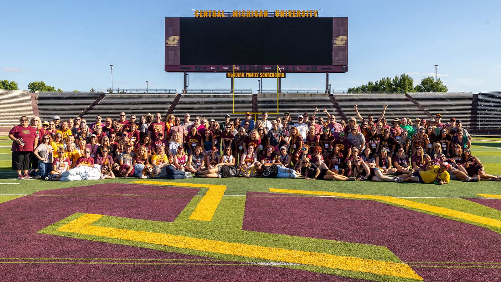 A large group of people pose for a photo on a football field in front of a jumbo scoreboard.