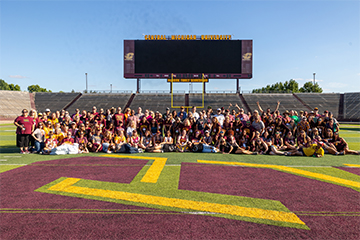 A large group of people pose for a photo on a football field in front of a jumbo scoreboard.