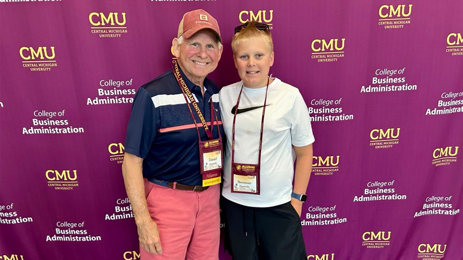 A senior citizen man and a younger boy pose for a picture in front of a Central Michigan University College of Business Administration step and repeat.