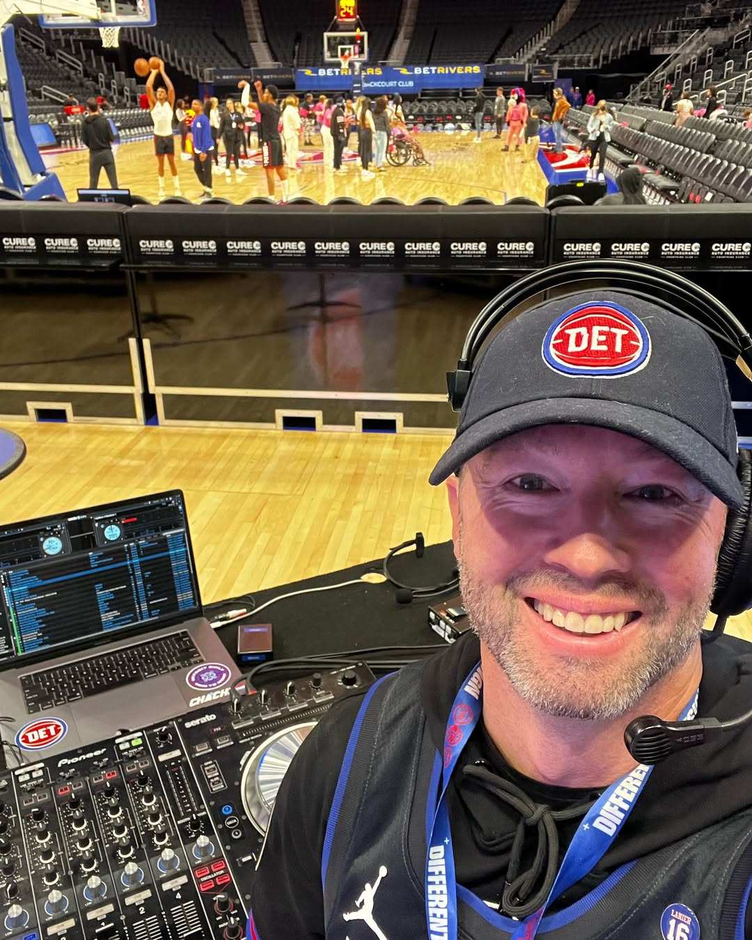 A man wearing headphones in front of a music mixer is smiling at camera with basketball players on a court behind him.