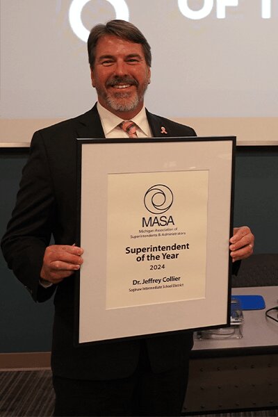 This is a photo of a man in a dark suit, white dress shirt, and striped pink tie holding a plaque sign that says MASA Superintendent of the Year 2024.