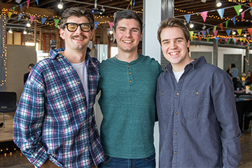 Three young men pose for a picture. Each has brown hair. The one on the left is wearing glasses and a plaid shirt. The one in the middle wears a green henley shirt, and the one on the right wears a navy blue long-sleeve shirt.