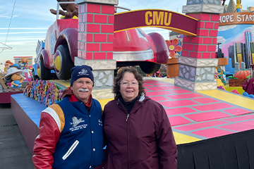 A man wearing a blue letterman's jacket and blue winter hat poses with a woman in a maroon zip-up coat in front of the CMU float at America's Thanksgiving Parade.