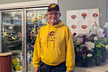A man in a yellow Central Michigan Alumni sweatshirt with a maroon baseball hat is surrounded by flowers inside a flower shop.
