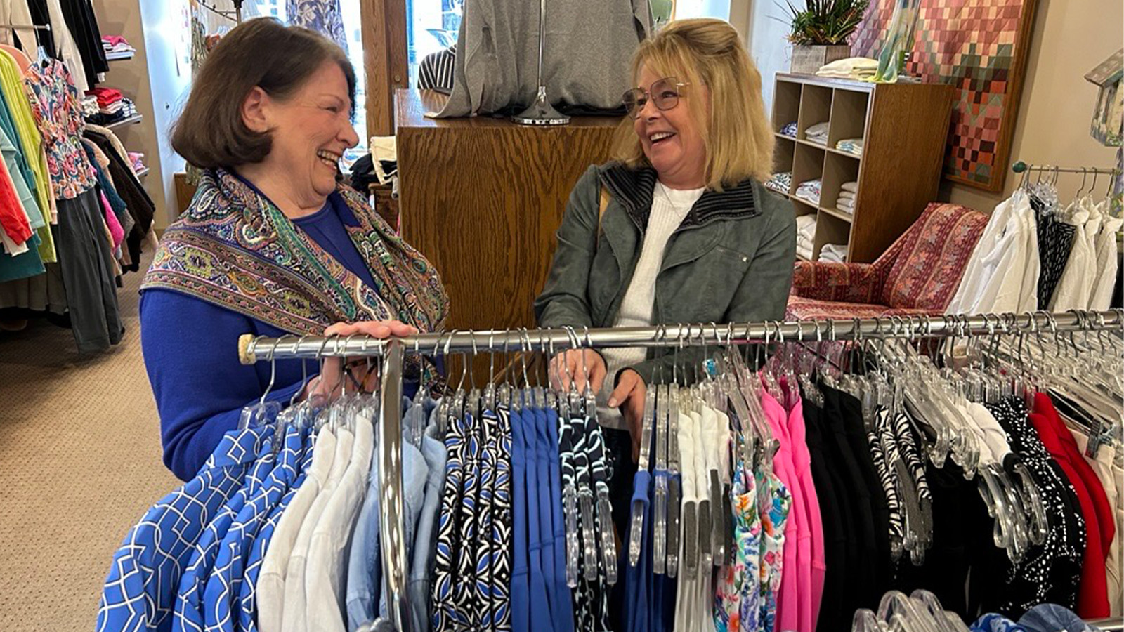 Two women smiling at each other behind a clothing rack.