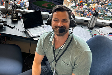 A man wearing headphones and a microphone poses for a photo while sitting in a stadium press box.