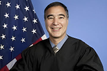 A man with short dark hair dressed in a black court robe and light-colored necktie poses in front of the US flag.