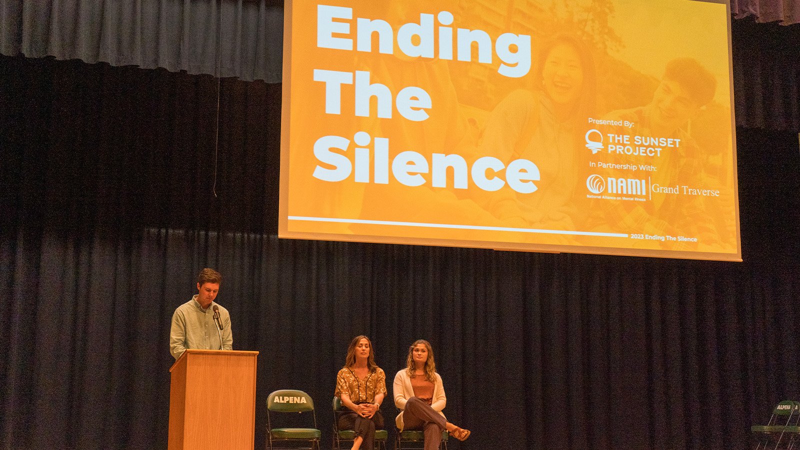 A young man wearing a light-colored shirt stands behind a wooden podium on a stage, and two females are sitting in a chair behind him. A sign that says Ending the Silence is hanging in the background.