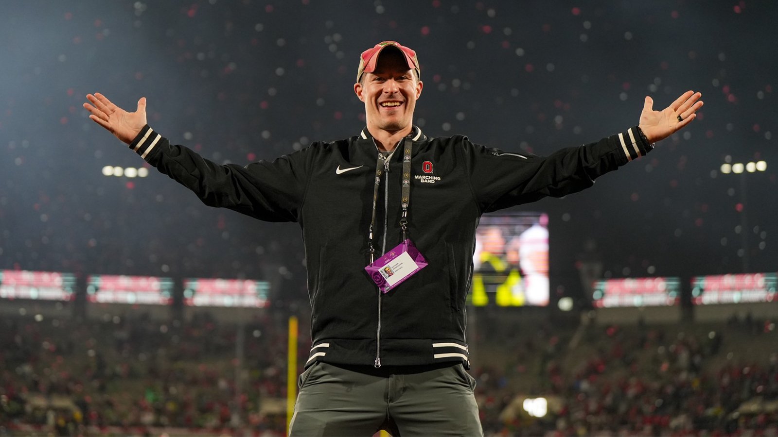 A man wearing a black zipped-up jacket and red hat stands and smiles with his arms outstretched.