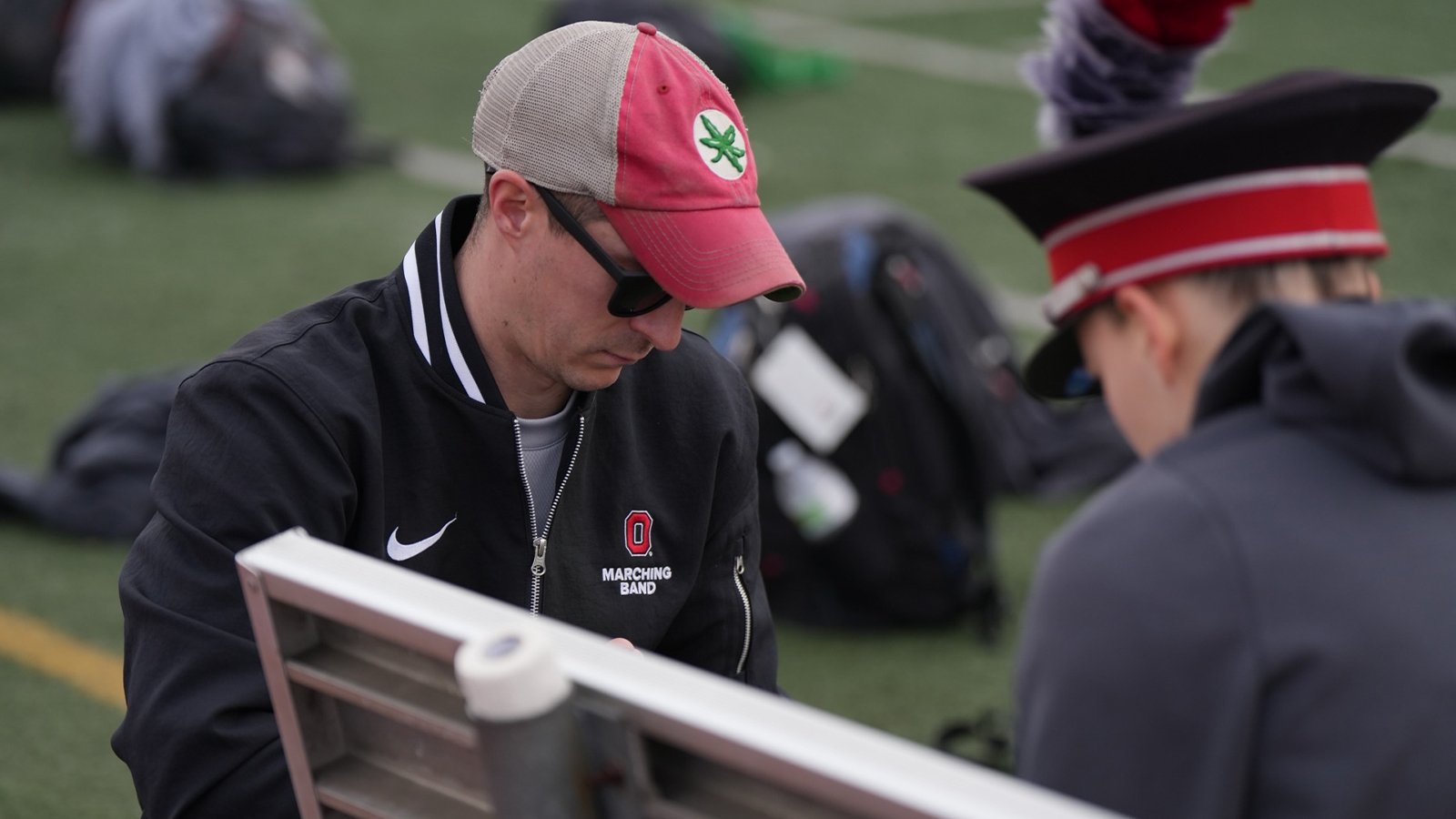 A man in a black jacket, red hat, and black sunglasses tends to a marching band member sitting on a bench.