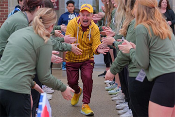 A man wearing a maroon and gold jumpsuit and ballcap happily gives high-fives while running through a human-formed tunnel of college students dressed in light green shirts.