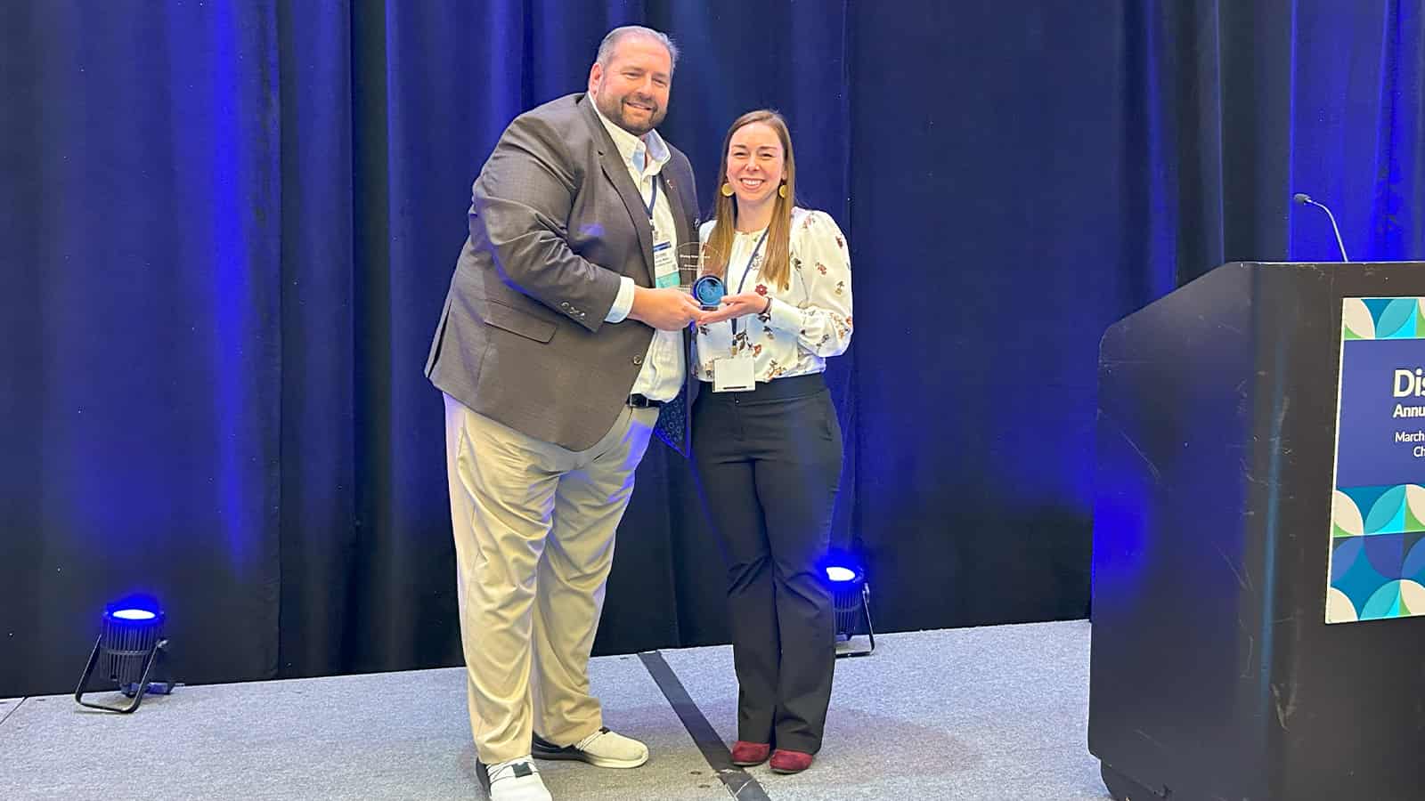 A bearded man wearing khaki pants and a suit jacket holds a small trophy with a brown-haired woman wearing dark slacks and a white long-sleeved shirt.