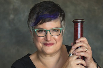 This is a headshot photo of a woman with a small portion of purple-dyed hair amidst a full head of dark hair, wearing teal-framed eyeglasses while holding a musical instrument.