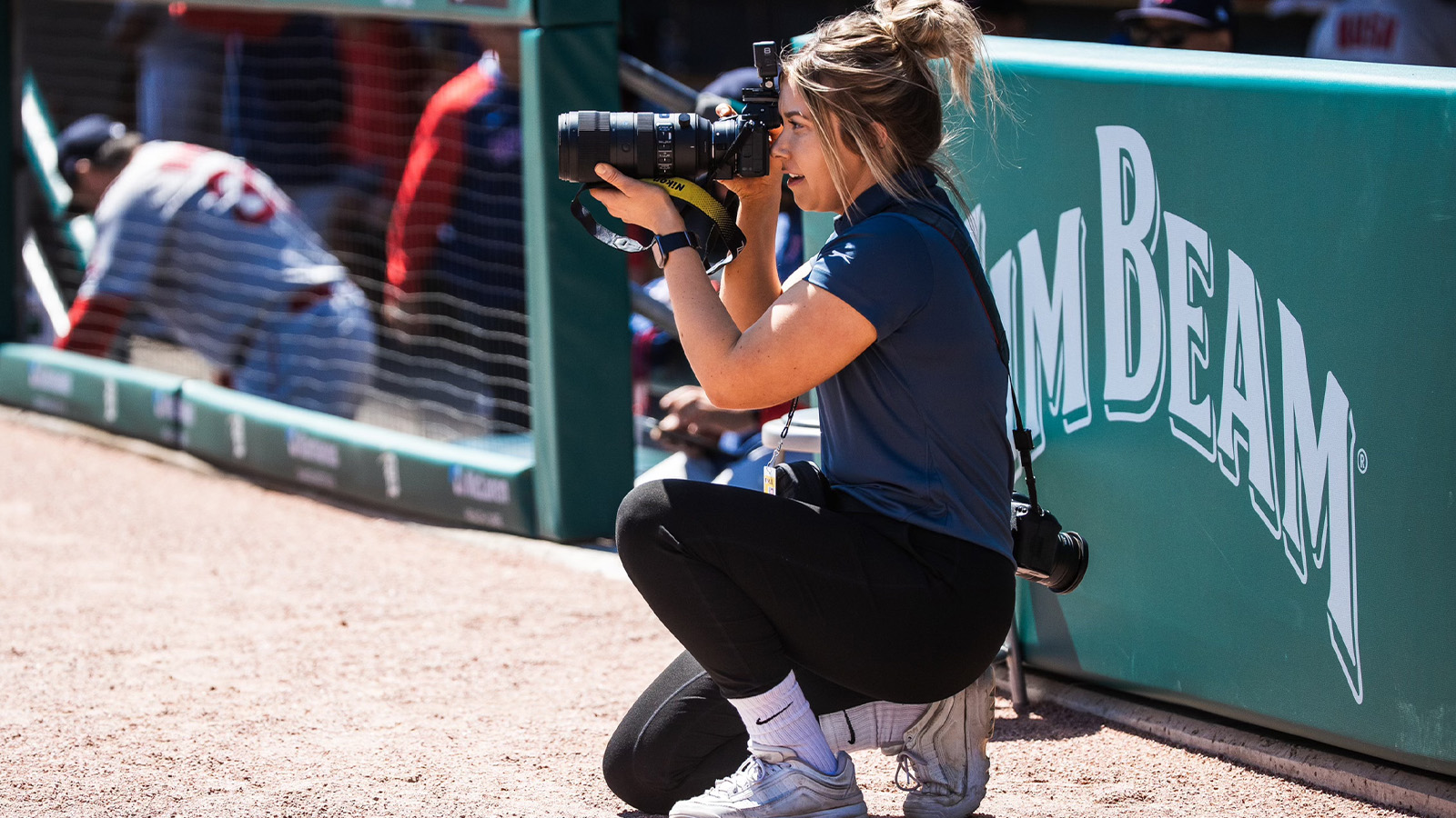 Woman with sandy brown hair wearing a blue shirt and black pants is crouching in front of a dugout with a camera in front of her face.