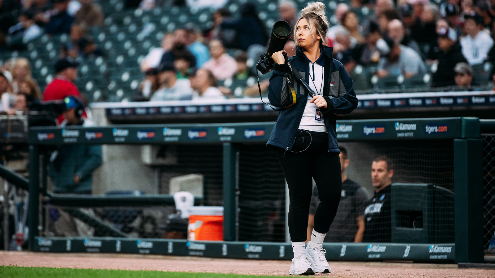 Woman with sandy brown hair wearing a jacket, white shirt, and black pants walks in front of a dugout while carrying a camera on a tripod.