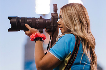 Woman with blond hair wearing a light blue shirt is holding and looking into a professional camera.