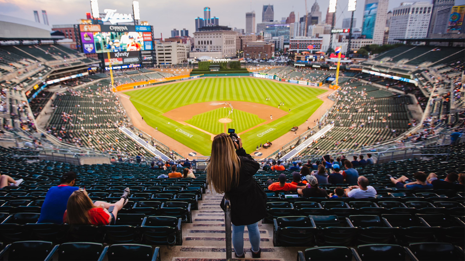 This is a panoramic picture from the upper deck level inside Comerica Park in Detroit, facing the ball field with a woman in the foreground holding a camera in front of her face.