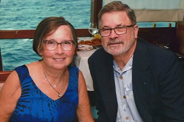 A man in glasses wearing a navy blue sports coat poses next to a woman in glasses wearing a blue and black dress.