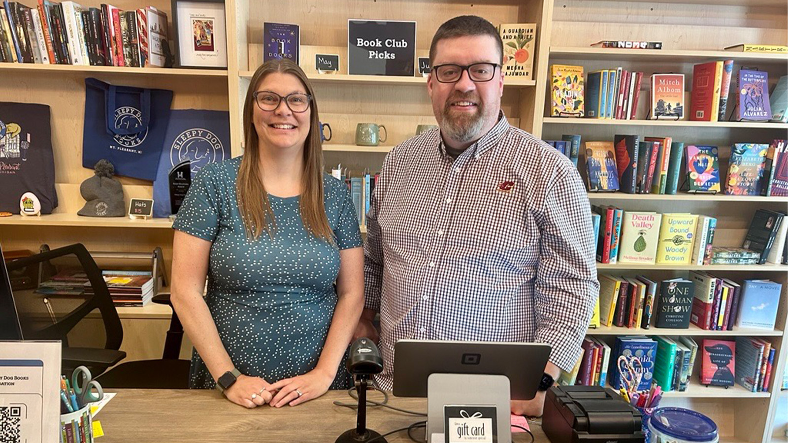 A man and woman standing in front of a computer with bookshelves filled with books behind them.
