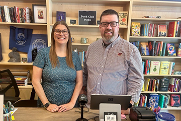 A man and woman standing in front of a computer with bookshelves filled with books behind them.