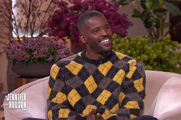 A man with short black hair and a trimmed beard is smiling wearing a black, yellow, and grey argyle sweater while being interviewed.