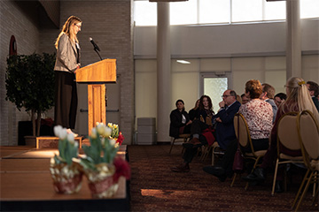 A young woman in dark pants and a long-sleeved light brown jacket stands behind a podium and speaks to a crowded room.