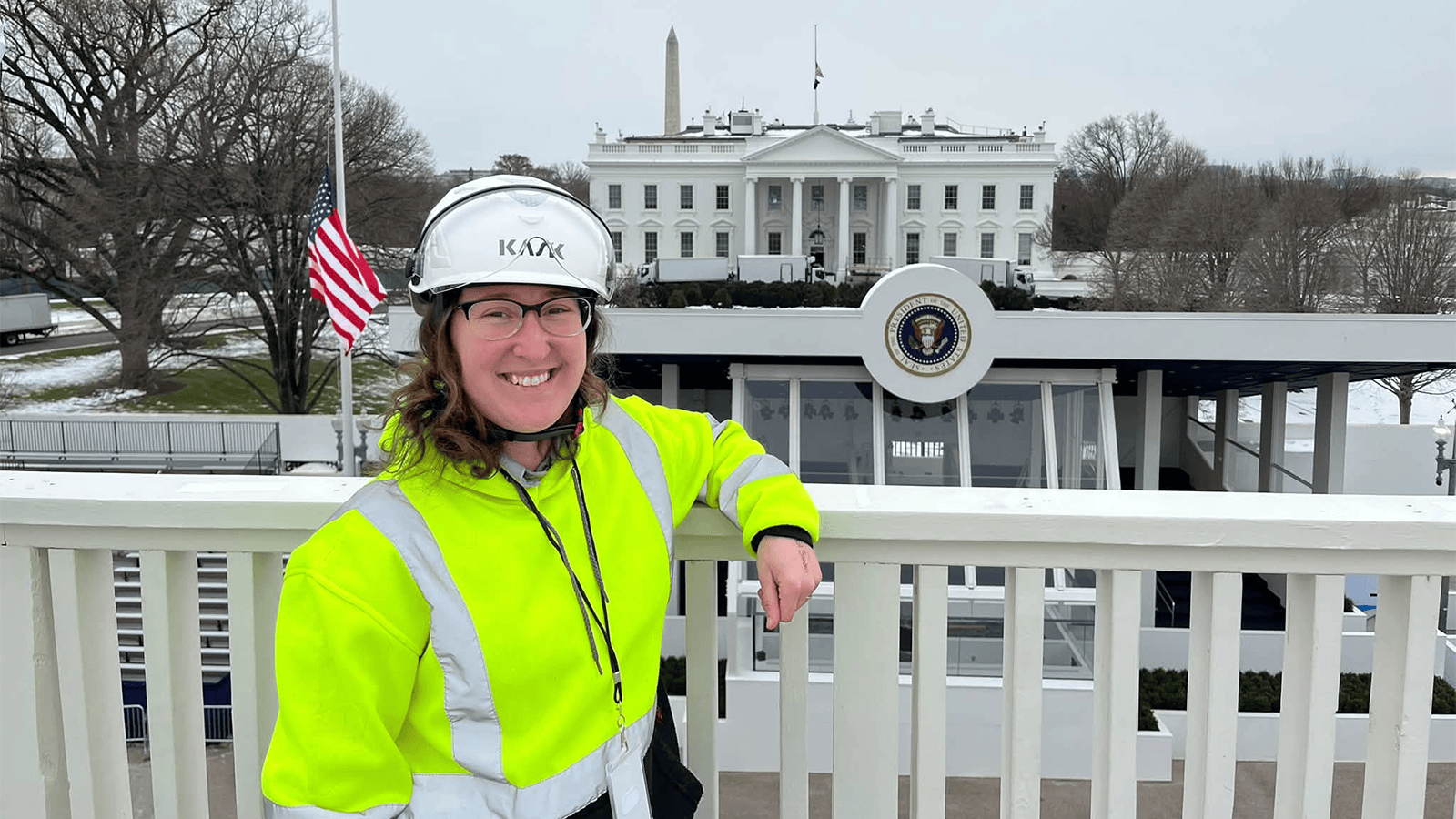 A woman wearing a yellow safety vest and a white helmet stands against a fence with The White House in the background.
