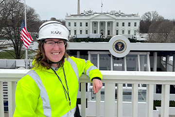 A woman wearing a yellow safety vest and a white helmet stands against a fence with The White House in the background.