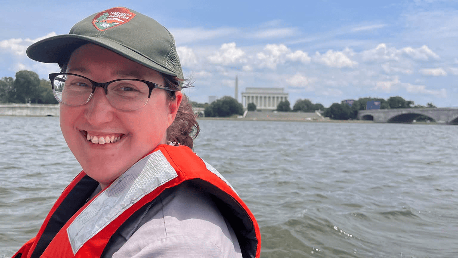 A woman wearing a hat and glasses is smiling at the camera with a body of water in the background.