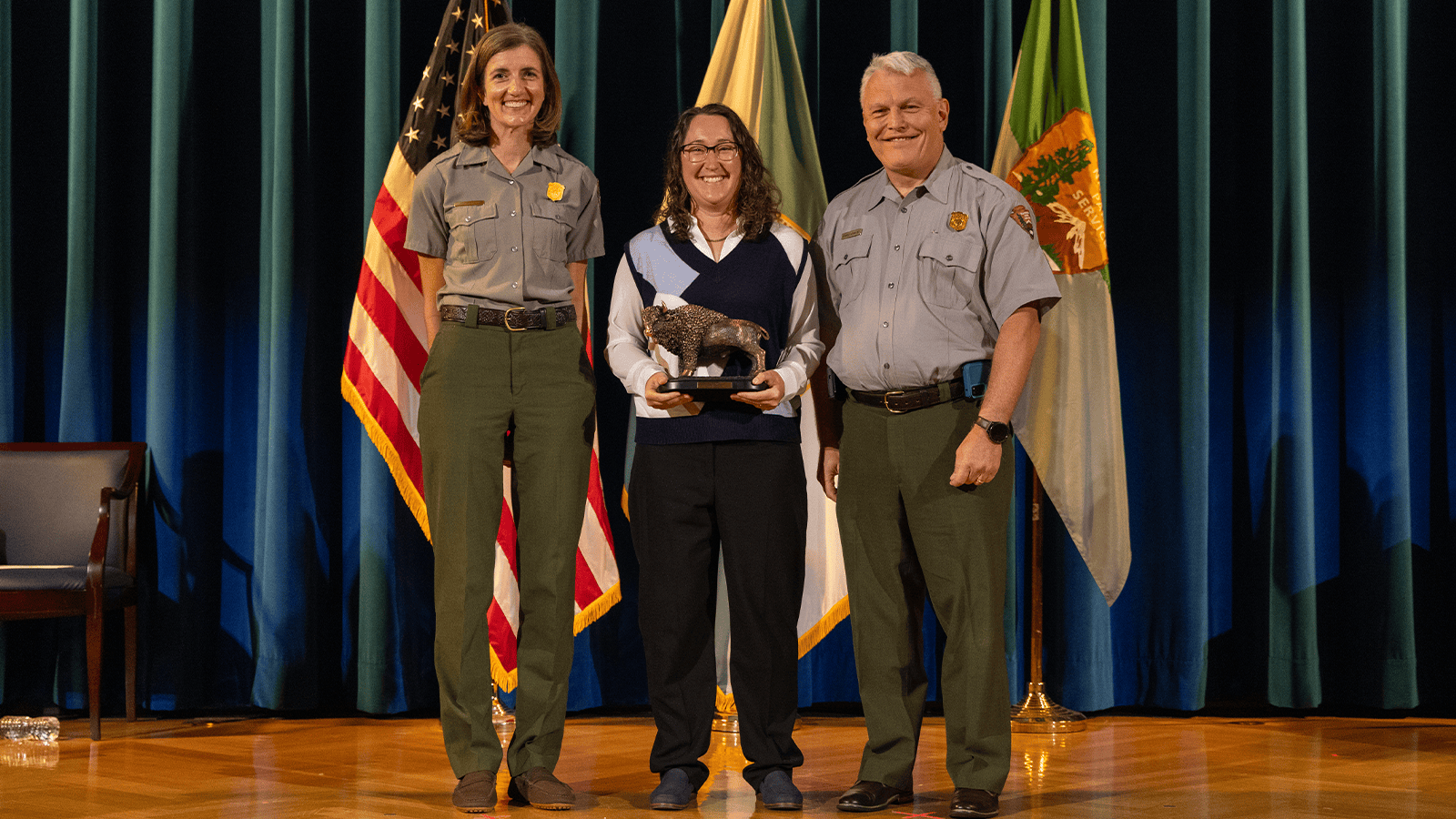 Two people in park ranger outfits stand on a stage while a woman in a sweater and black pants holding an award stands between them.