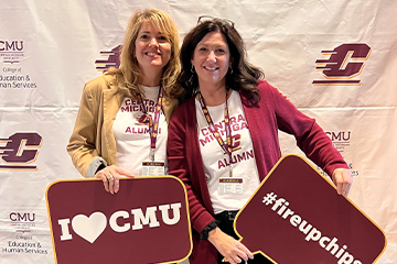 Alumnae pose in front of a CMU logo wall. Both are wearing white Central Michigan alumni shirts under blazers. One wears a gold blazer holding a sign that says I heart CMU and the other wears a maroon blazer holding a sign that says fireupchips.