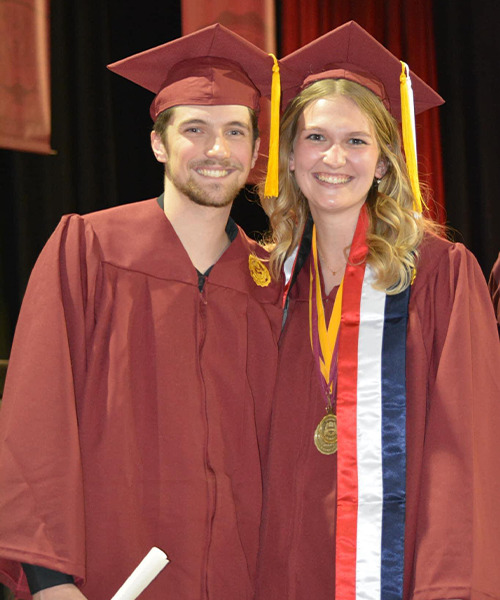 A male and female smile and pose together after a commencement ceremony, each wearing a cap and gown.