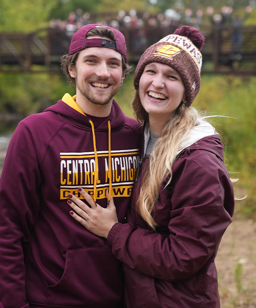 A man wearing a CMU hooded sweatshirt and backward ballcap smiles and poses with a woman wearing a maroon jacket and CMU winter hat while showing off her engagement ring.