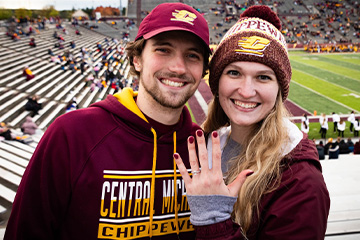 A man wearing a CMU hooded sweatshirt and action C hat smiles and poses alongside a woman who is smiling while wearing a CMU winter hat and showing off her engagement ring inside Kelly/Shorts Stadium.