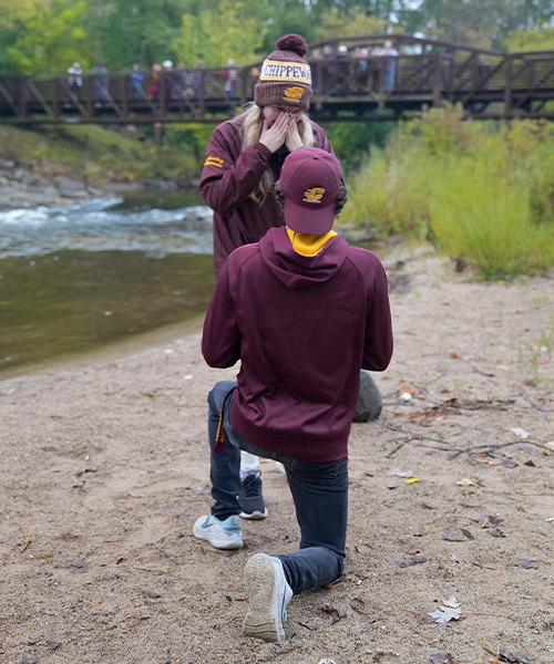 A man wearing a maroon hooded sweatshirt and backward action C ballcap is down on one knee in the sand beside a river, proposing to a woman wearing a maroon jacket and CMU winter hat.