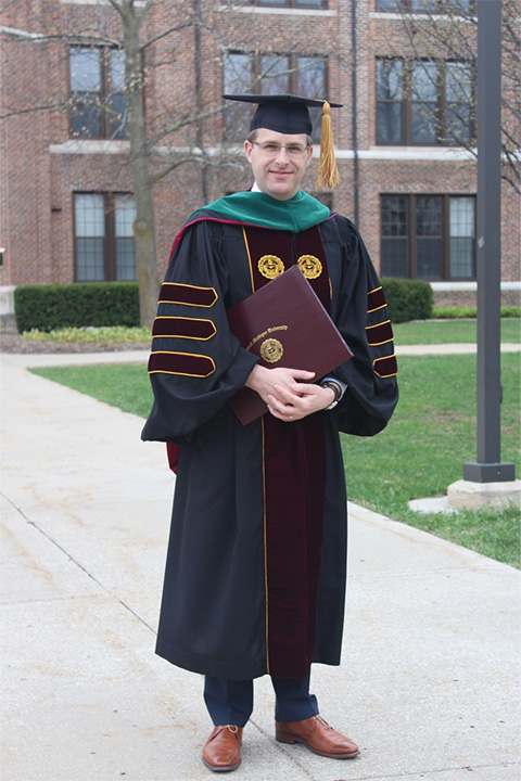 A man in a graduation cap and gown is holding his degree on a sidewalk in front of a building.