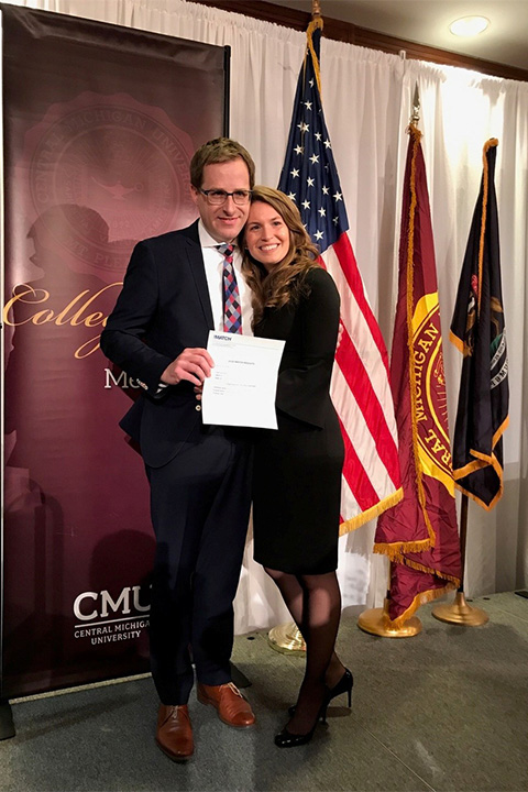 A woman has her arms around a man who is holding a piece of paper in front of the US flag, CMU flag and state of Michigan flag.