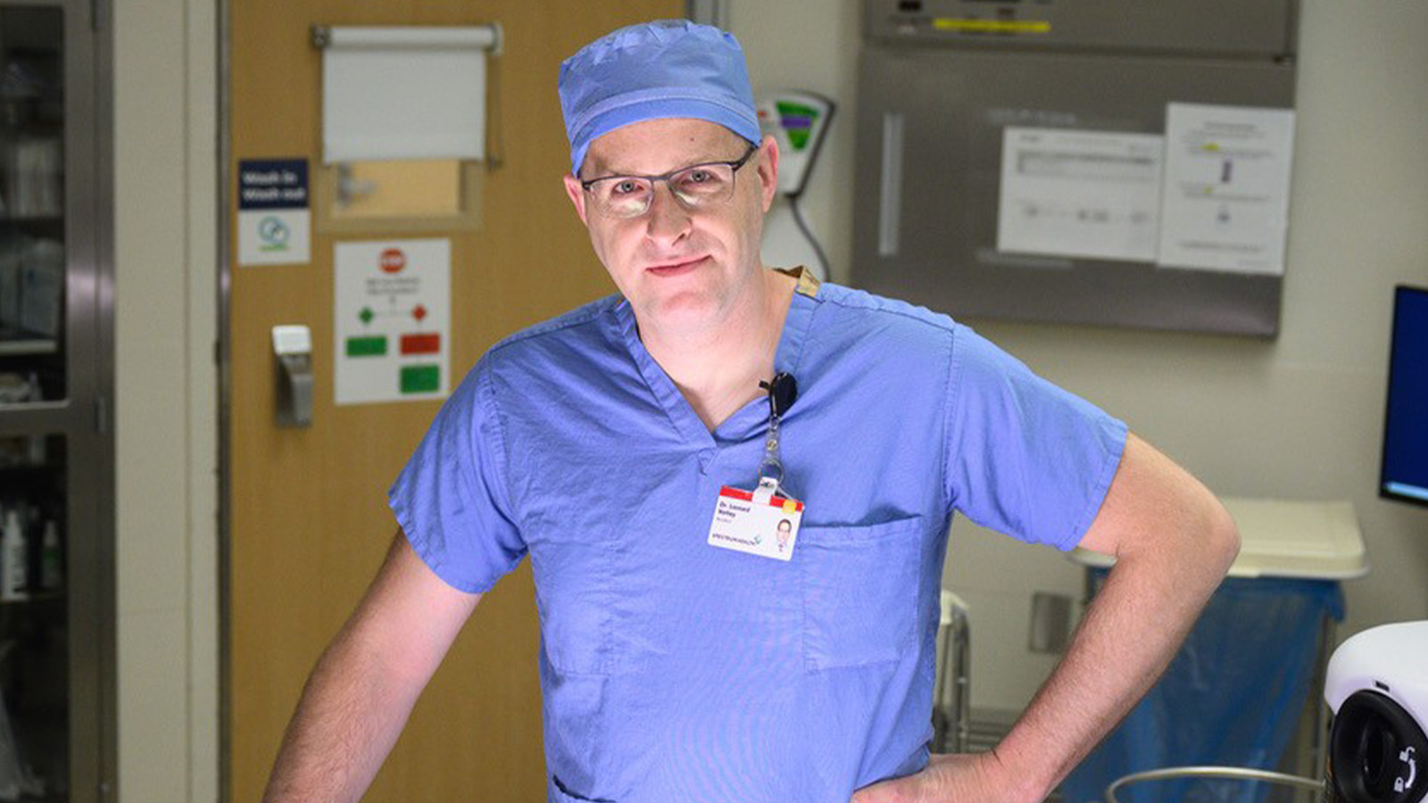 A man with glasses on is wearing scrubs inside a hospital room.
