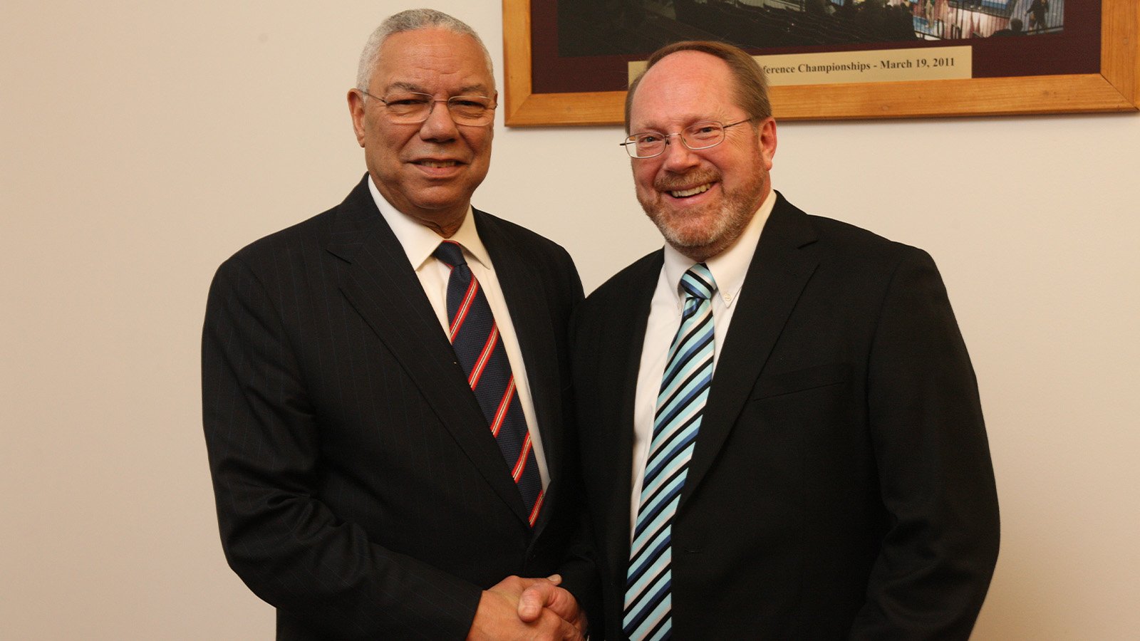 Two men wearing suits with striped ties shake hands in front of a light brown wall.