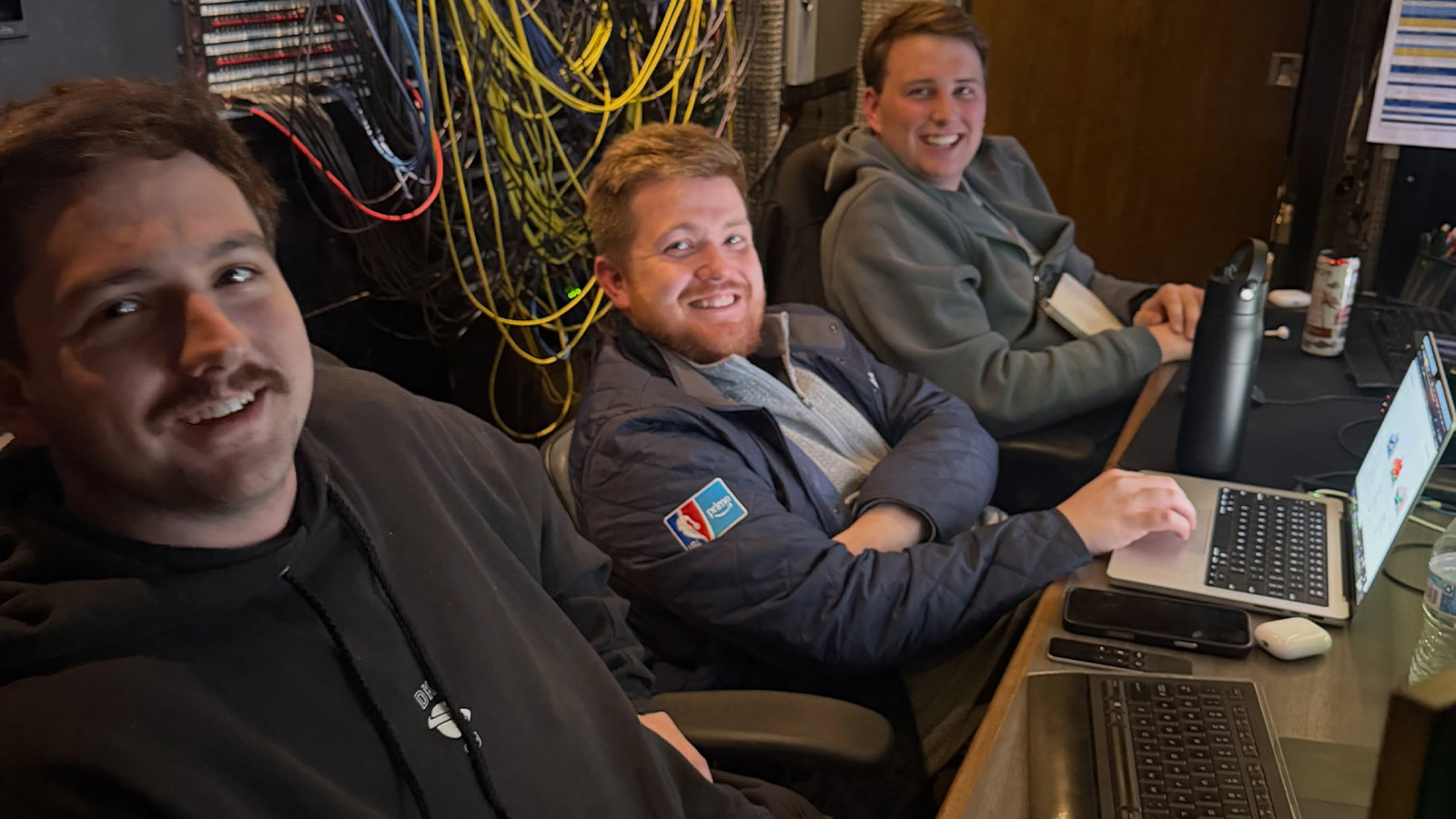 A group of men sitting at a desk with computers and many cords hanging on the wall behind them.