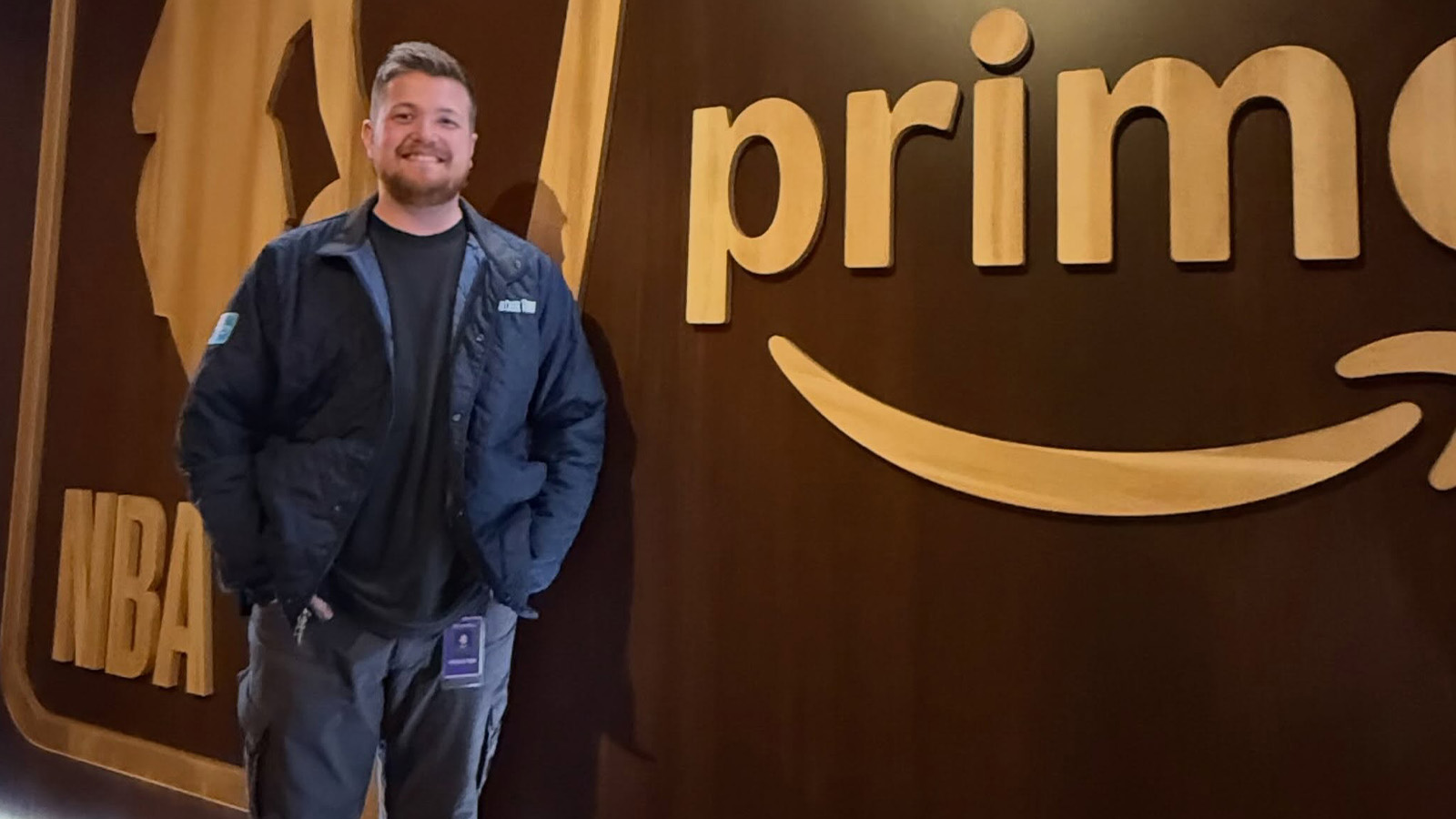 Zach Newhouse next to NBA on Prime wall 1600x900 A man in a navy jacket stands in front of a wall that has