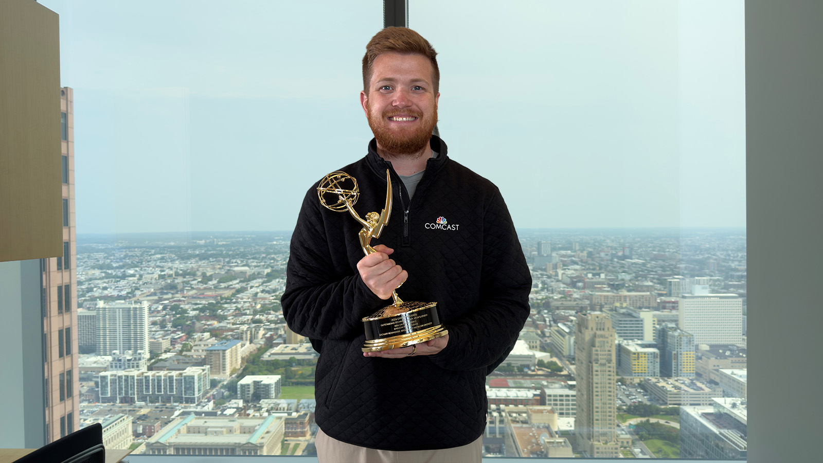 Zach Newhouse with Emmy in front of cityscape window 1600x900 A man with a beard wearing a black quarter zip holds a trophy in front of a window with the view of a city in the background.