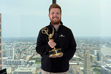 A man with a beard wearing a black quarter zip holds a trophy in front of a window with the view of a city in the background.