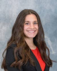 Profile portrait for Morgan Lyon, Counselor Intern for the Counseling Center. She has long brown hair, wearing a red shirt and a black blazer. The background is blue and grey.