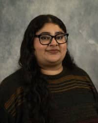 Muskan Sany, COMPASS Peer Educator. Dark long hair, glasses, wearing a dark shirt with stripes in front of a grey background.