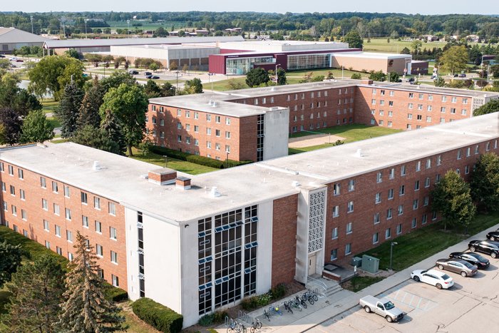 saxe-housing-int-nav Aerial view of Saxe Hall on the campus of Central Michigan University, captured from a drone. The residence hall is surrounded by lush green grass and trees, with clear blue skies above.