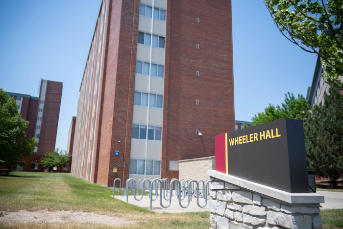 Wheeler Hall sign in front of a tall red brick residence building with vertical windows and bike racks nearby.