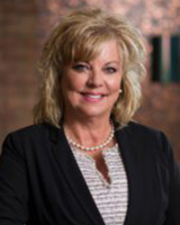 Woman with blonde hair, wearing black suit jacket smiling against brown background.