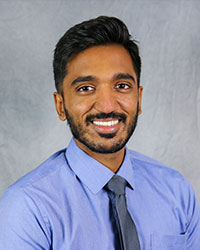 A man with styled dark short hair and facial hair, smiles for a headshot photo wearing a blue shirt and tie.